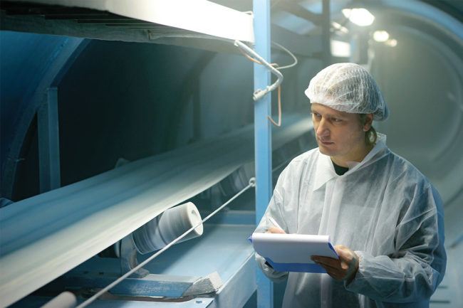 Man in white clean suit evaluating industrial processing machinery (©STOCKR - STOCK.ADOBE.COM)